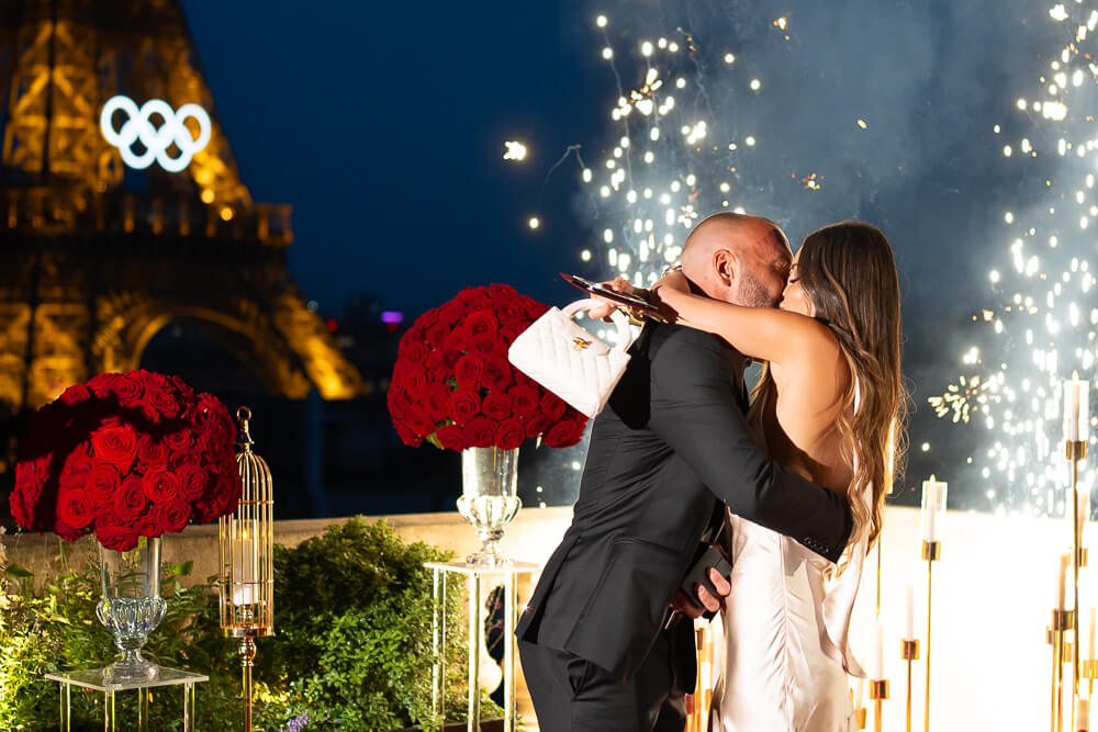 Couple kissing on an exclusive Eiffel Tower terrace at Shangri-La Hotel Paris, surrounded by fireworks and vibrant red roses under a vivid blue sky.