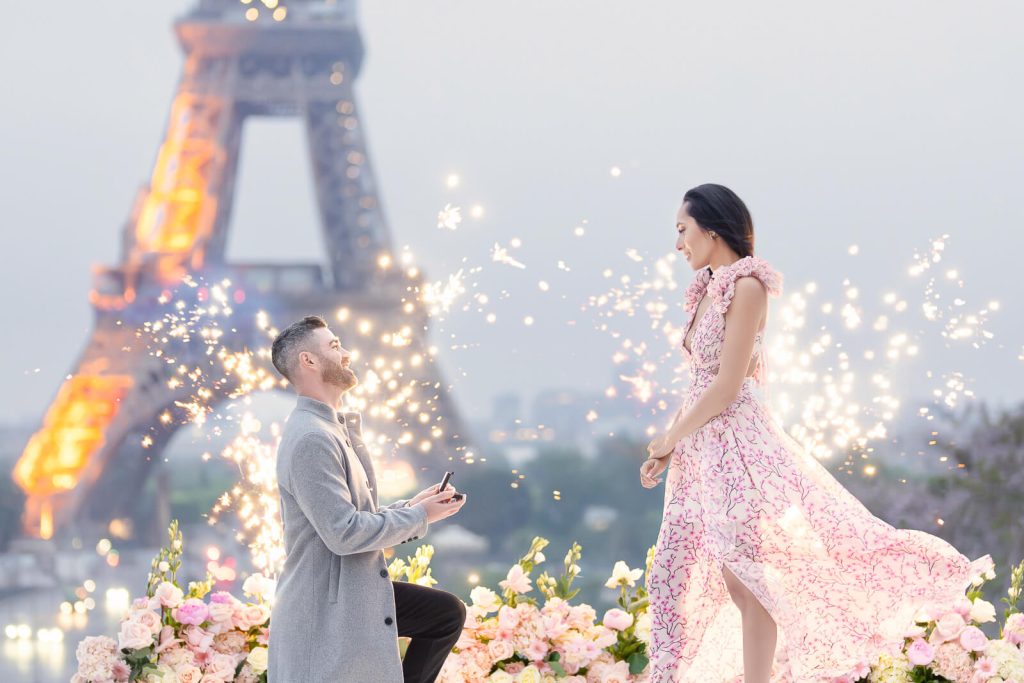 Enlisting a seasoned local Paris proposal planner is key to navigating the complexities of public hotspots like Trocadéro, as seen in this legendary Eiffel Tower proposal. Here, a stylish couple steals the show as the gentleman drops to one knee, presenting a ring box to his sweetheart, surrounded by stunning florals and sparkling fireworks—a truly magical ambiance.