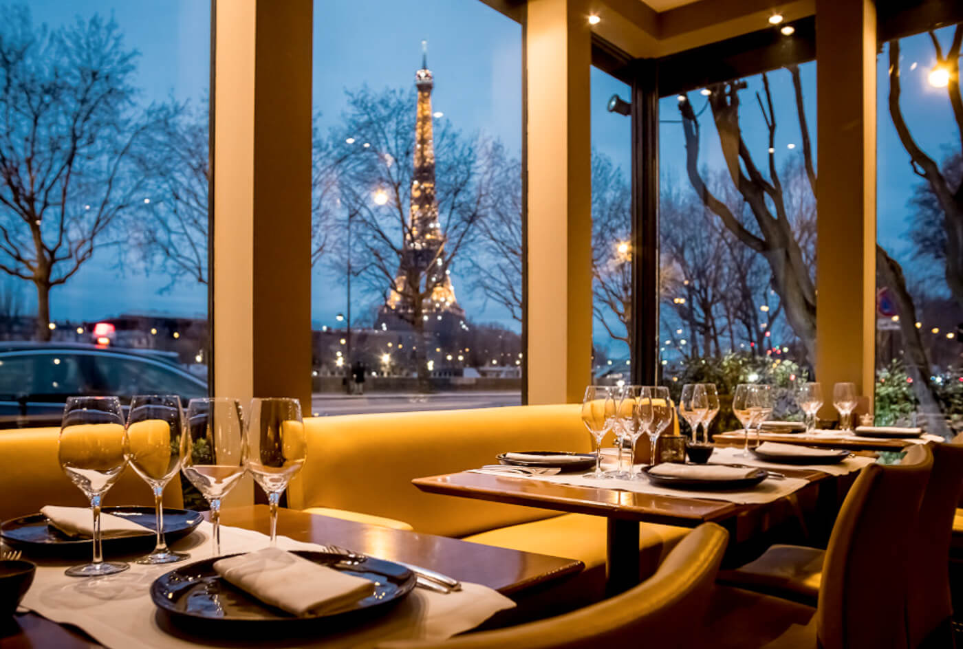6 New York restaurant's warmly lit dining room with towering windows framing the illuminated Eiffel Tower against a deep blue twilight sky