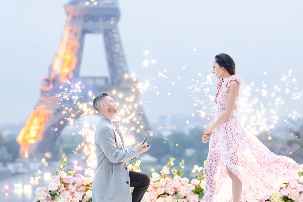 Couple at Trocadéro Plaza at sunrise, man kneeling with ring box on a white carpet and pink rose-petal aisle, surrounded by candles and sparkler fountains, Eiffel Tower illuminated in the background, bride-to-be in pink dress.