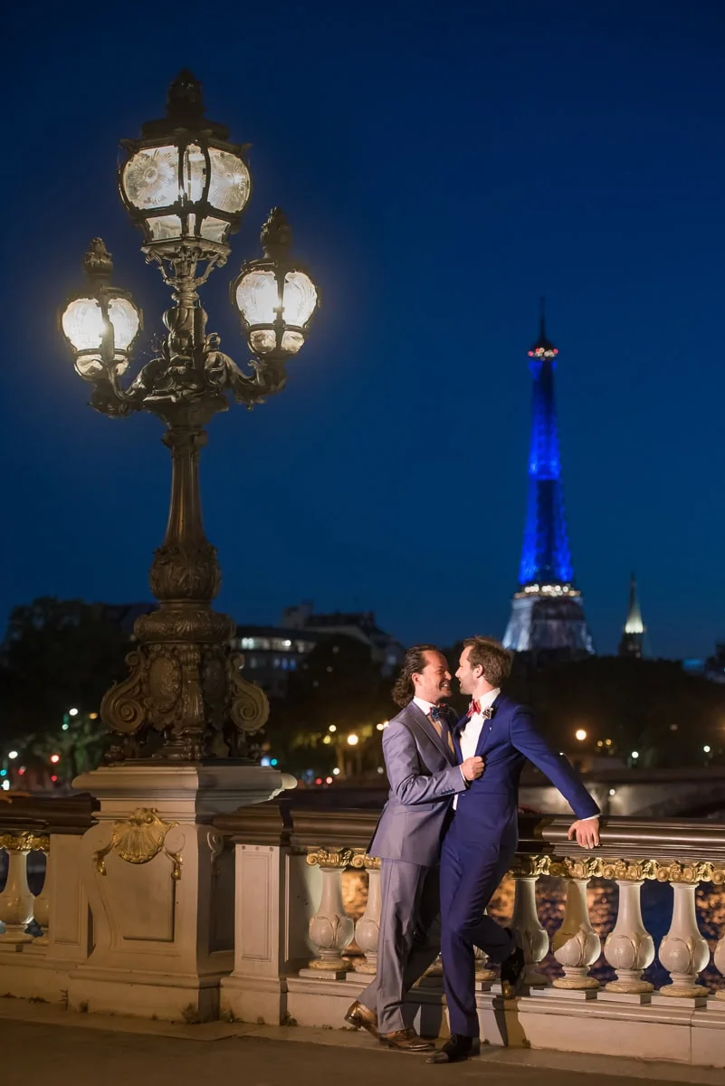 Same-sex male couple connecting playfully on Pont Alexandre III at night with blue-lit Eiffel Tower and ornate lantern lighting, Paris