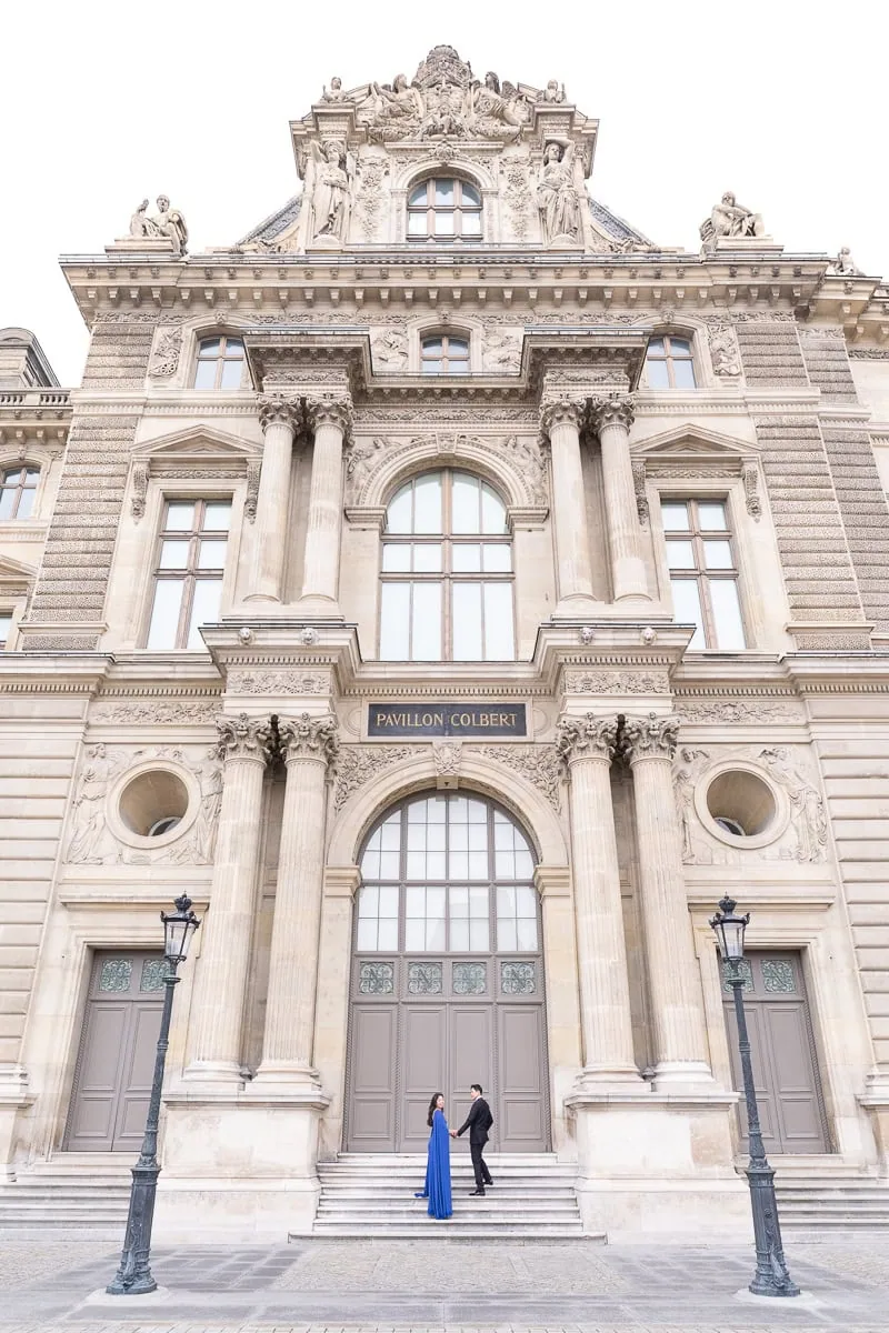 Couple ascending Colbert Pavilion staircase at Louvre Museum, she in blue gown looking back at camera while he leads her by hand against grand stone architecture