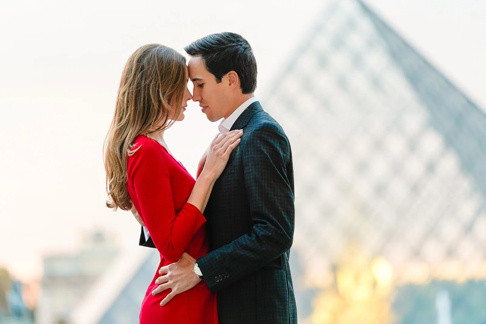 Romantic couple forehead to forehead at Louvre Museum during golden hour with pyramid softly blurred in background