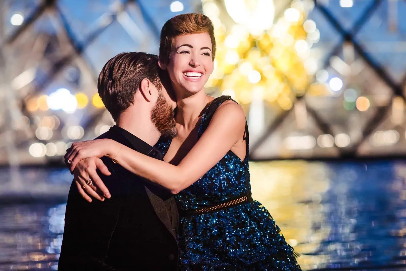 Joyful engaged couple laughing at Louvre Pyramid with fountain reflections during Paris engagement session