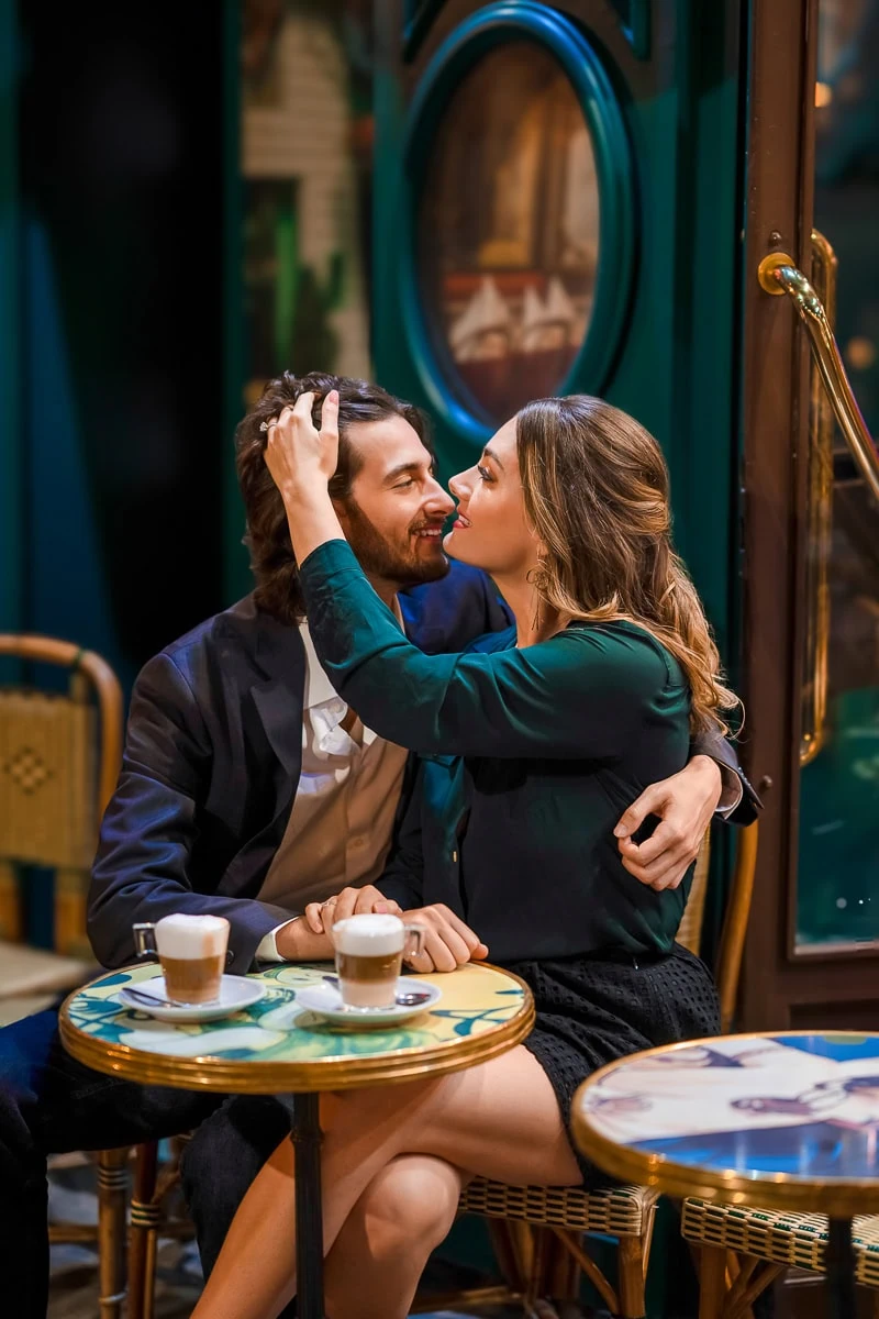 A couple shares a playful moment at a cafe table at night in Place du Tertre, Montmartre, illuminated by soft, continuous professional lighting.