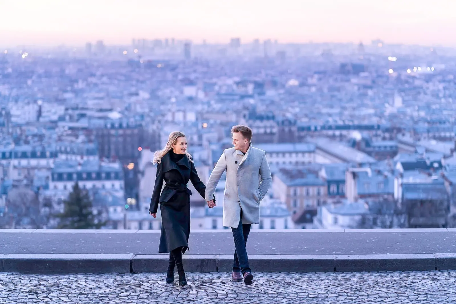 A couple holds hands walking towards the camera at sunrise from the panoramic viewpoint below the Sacré-Cœur Basilica in Montmartre, with a pink and blue sky.