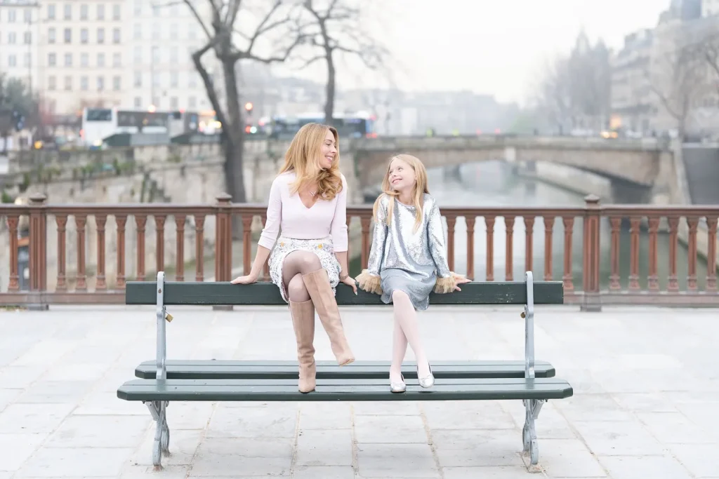 Paris family photographer captures mother and daughter sharing bench moment at sunrise near Notre Dame with Seine river backdrop - vacation family photography