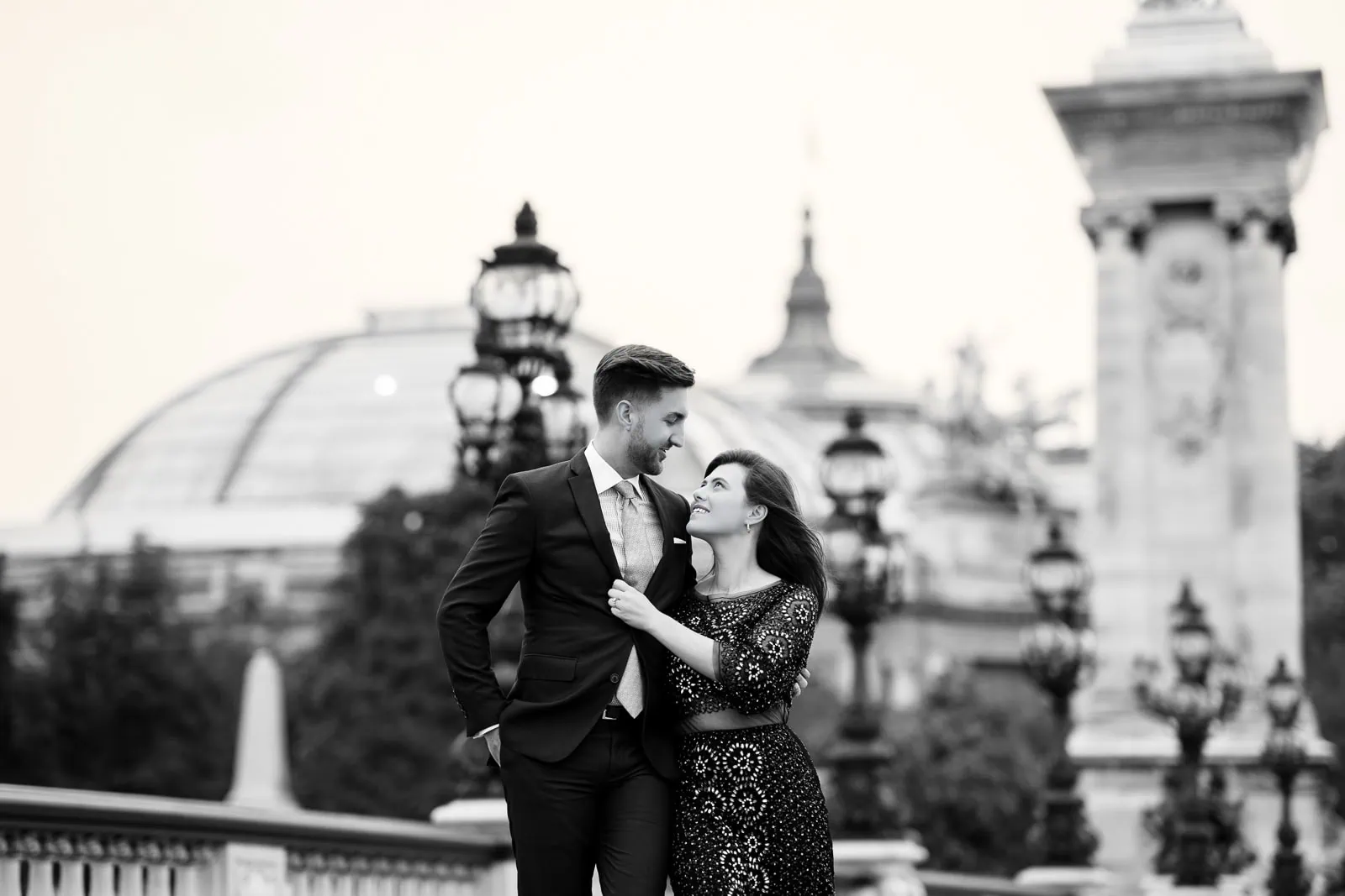 Stylish couple walking arm in arm in black and white on Pont Alexandre III, lanterns glowing overhead and the Grand Palais façade behind them