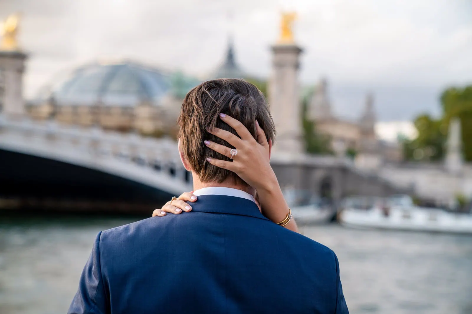 Engaged couple on Seine riverbank by Pont Alexandre III, bride's hands around groom's head highlighting diamond ring, with Grand Palais and bridge softly blurred