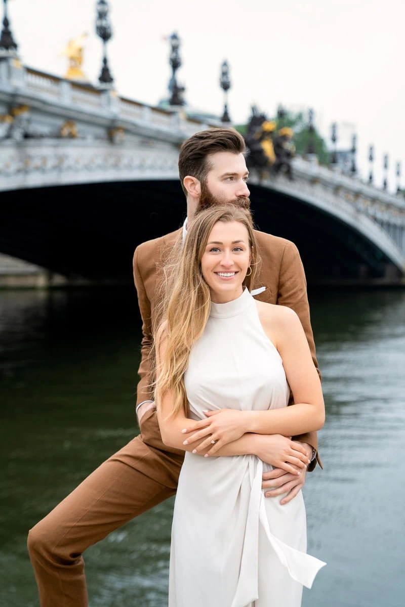 An elegant couple poses for a photograph on the cobblestone banks of the Seine River, with the arches of Pont Alexandre III above them in Paris.
