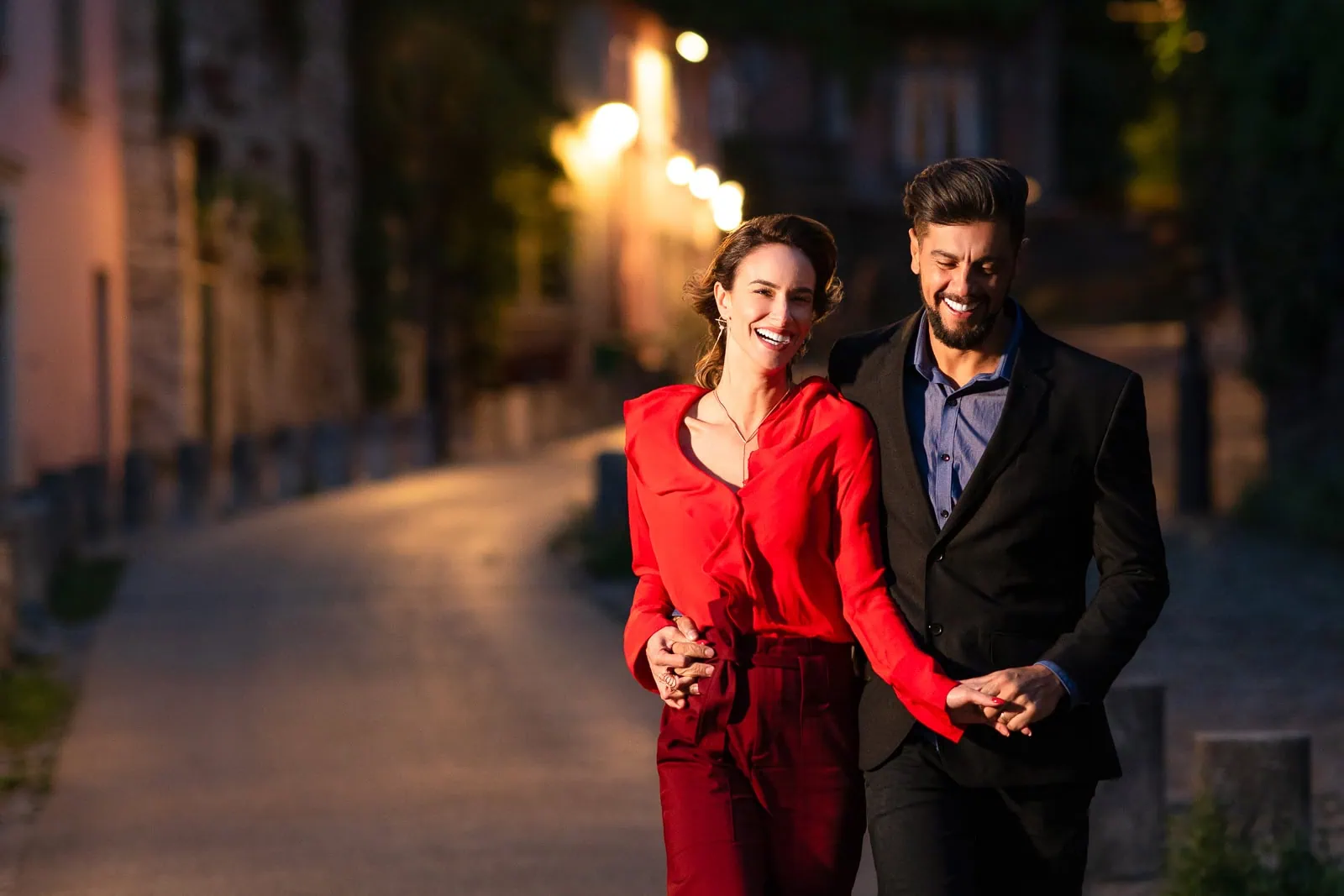 A joyful couple walks hand-in-hand at night on the winding Rue de l'Abreuvoir in Montmartre, with La Maison Rose visible uphill and Sacré-Cœur Basilica in the distance.