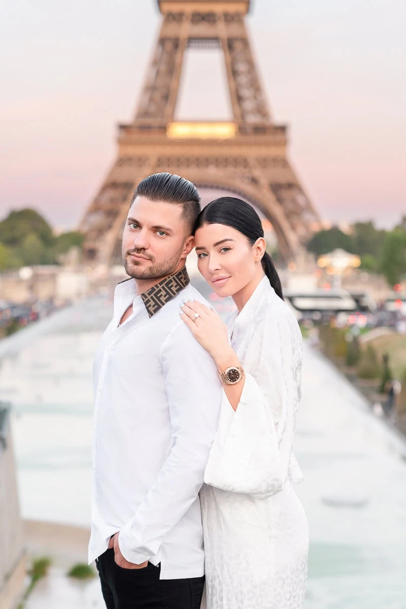 A couple poses for an evening engagement photo by the Warsaw Fountain at Trocadéro, with the woman displaying her new diamond ring.