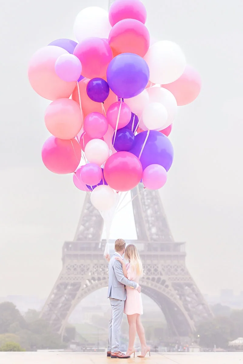 A couple faces the Eiffel Tower at sunrise from the left Trocadéro platform, holding an exceptionally large bouquet of balloons.