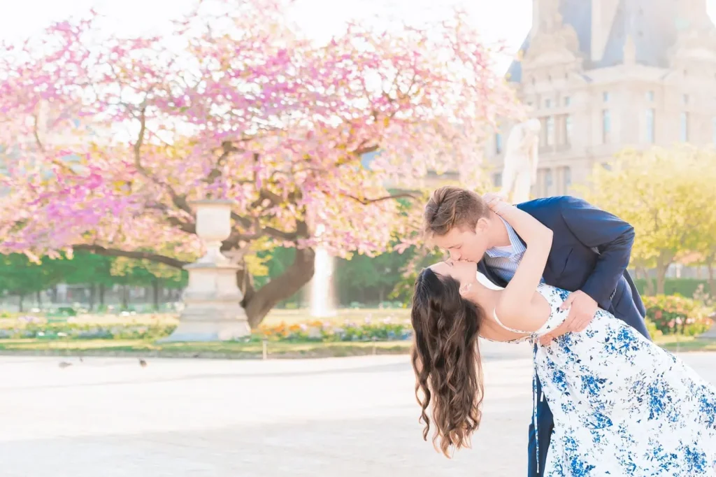Engaged couple sharing a dip kiss at sunrise amid cherry blossoms in Tuileries Garden, with the Louvre softly blurred behind them