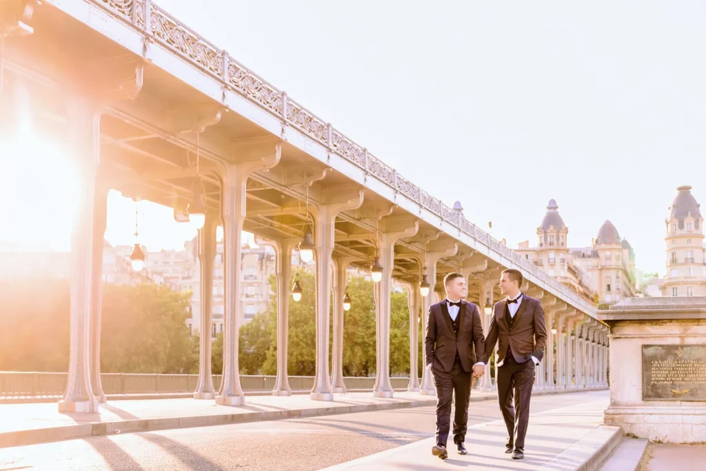 Same-sex couple in tuxedos holding hands walking Bir Hakeim Bridge during golden hour, Paris LGBTQ wedding photographer