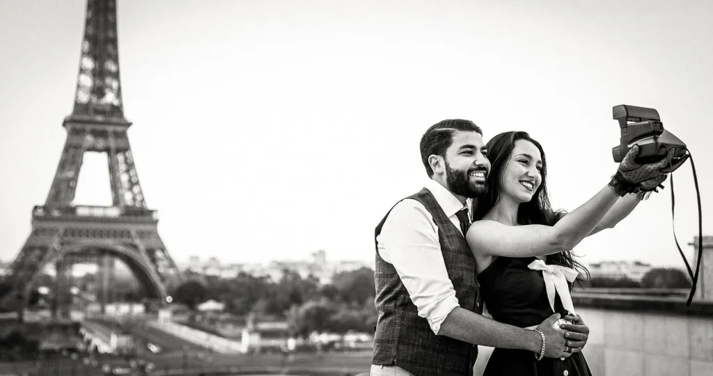 Couple in vintage outfits taking Polaroid selfie with Eiffel Tower backdrop, Charlie Chaplin themed black and white Paris wedding photographer