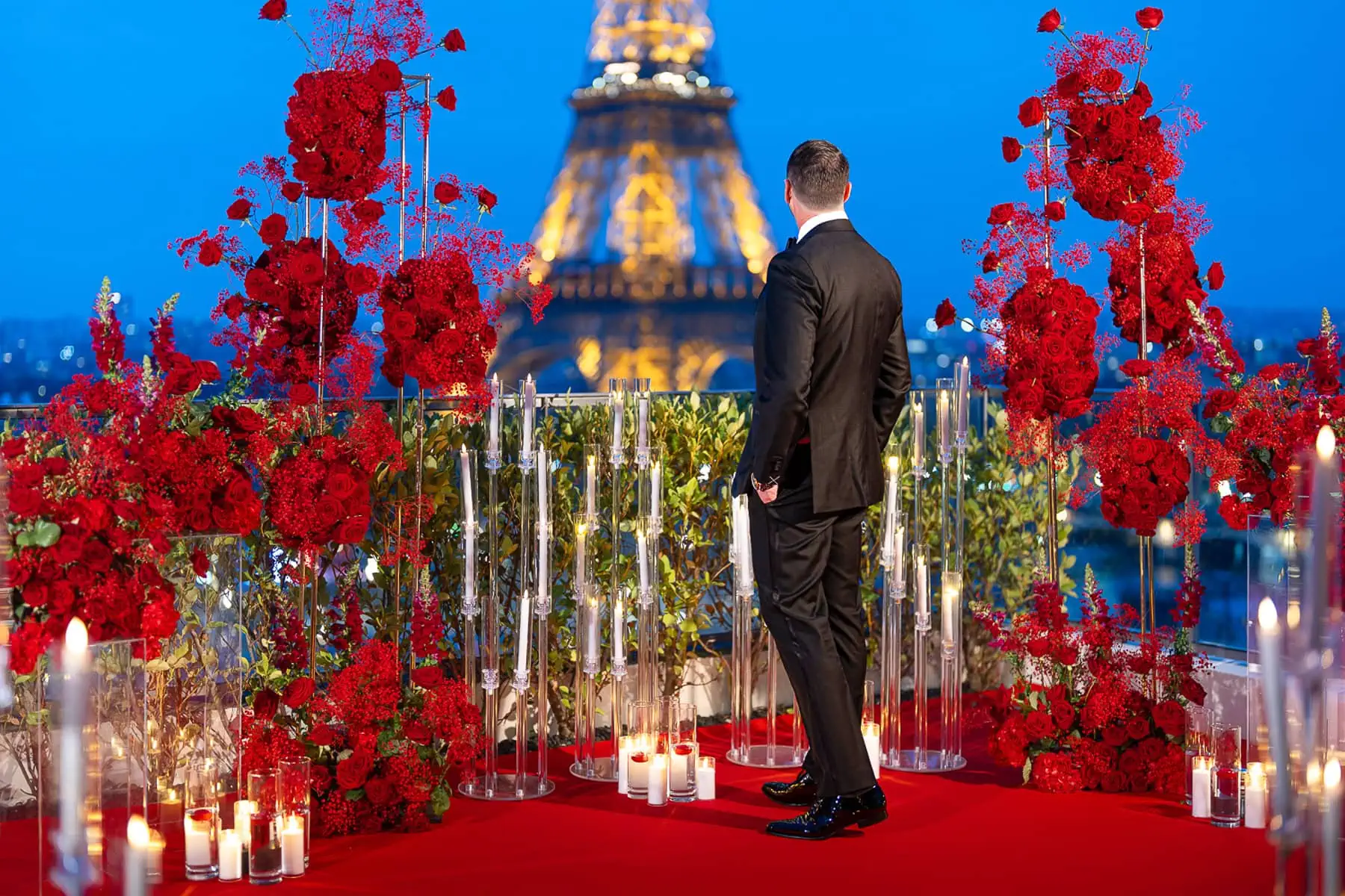 Blue hour Paris proposal at Shangri La Eiffel Tower penthouse terrace with man in tuxedo, red rose arrangements, candles, and illuminated tower.
